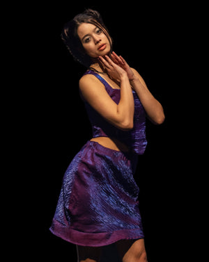 Model wearing a purple dress on a dark runway with an audience.